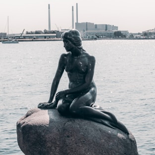 statue of woman sitting on rock near body of water during daytime
