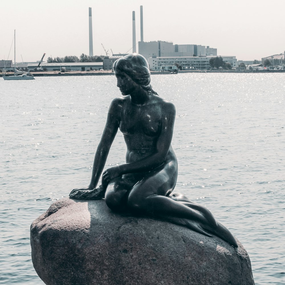 statue of woman sitting on rock near body of water during daytime