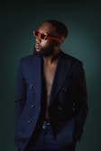 A well-dressed man wearing a navy blue double-breasted blazer with a bare chest. He sports red sunglasses and has a neatly groomed beard and styled hair. The background is dark green, creating a stylish and sophisticated atmosphere.
