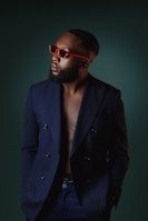 A well-dressed man wearing a navy blue double-breasted blazer with a bare chest. He sports red sunglasses and has a neatly groomed beard and styled hair. The background is dark green, creating a stylish and sophisticated atmosphere.