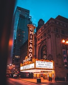 A brightly lit theater marquee in an urban setting displays event information. The marquee features classic vintage signage adorned with lights, set against the backdrop of tall city buildings. The street is dimly lit, suggesting evening or nighttime, and a few people are visible walking nearby.