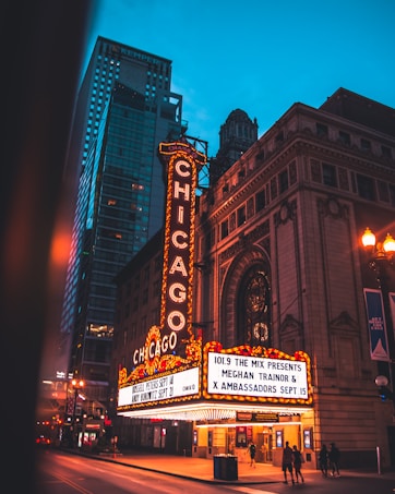 A brightly lit theater marquee in an urban setting displays event information. The marquee features classic vintage signage adorned with lights, set against the backdrop of tall city buildings. The street is dimly lit, suggesting evening or nighttime, and a few people are visible walking nearby.