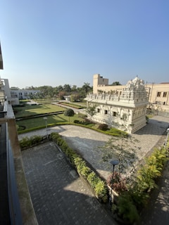 A well-maintained garden with a paved walkway and manicured hedges surrounds a white stone temple with intricate carvings. The temple is situated in a landscaped area with green grass, palm trees, and various plants. Modern buildings are visible in the background under a clear blue sky.