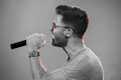 A man with sunglasses and a wristwatch is passionately singing into a microphone. He is captured in profile, showcasing his expression of intensity and engagement. The background is blurred, emphasizing the focus on the performer.