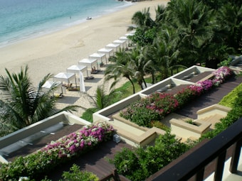 A beautiful beachfront scene featuring a row of white cabanas lined up along the sandy shore, bordered by lush palm trees. The foreground showcases a terraced area filled with vibrant pink and white flowers and well-maintained greenery. The ocean is calm, with gentle waves lapping the shore, and a few people can be seen enjoying the beach in the distance.