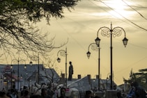 An evening scene with silhouetted people and horses in front of antique street lamps. The background features a tall statue, architectural buildings, and a tree with branches stretching across the sky. The sky has a warm, golden hue as the sun begins to set.