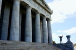 A person stands triumphantly on the steps of a large, classical-style building with tall stone columns. The sky is bright and clear, adding to the monumental feeling of the scene.