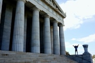 A person stands triumphantly on the steps of a large, classical-style building with tall stone columns. The sky is bright and clear, adding to the monumental feeling of the scene.