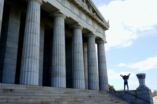 A person stands triumphantly on the steps of a large, classical-style building with tall stone columns. The sky is bright and clear, adding to the monumental feeling of the scene.