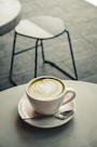 A white cup of matcha latte with intricate latte art sits on a matching saucer. A small spoon rests on the saucer beside the cup, which is placed on a round, light-colored table. In the background, a blurred metal chair is visible on a tiled floor.