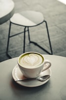 A white cup of matcha latte with intricate latte art sits on a matching saucer. A small spoon rests on the saucer beside the cup, which is placed on a round, light-colored table. In the background, a blurred metal chair is visible on a tiled floor.