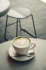 A white cup of matcha latte with intricate latte art sits on a matching saucer. A small spoon rests on the saucer beside the cup, which is placed on a round, light-colored table. In the background, a blurred metal chair is visible on a tiled floor.