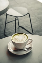 A white cup of matcha latte with intricate latte art sits on a matching saucer. A small spoon rests on the saucer beside the cup, which is placed on a round, light-colored table. In the background, a blurred metal chair is visible on a tiled floor.