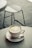 A white cup of matcha latte with intricate latte art sits on a matching saucer. A small spoon rests on the saucer beside the cup, which is placed on a round, light-colored table. In the background, a blurred metal chair is visible on a tiled floor.