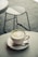 A white cup of matcha latte with intricate latte art sits on a matching saucer. A small spoon rests on the saucer beside the cup, which is placed on a round, light-colored table. In the background, a blurred metal chair is visible on a tiled floor.