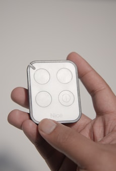 A hand holding a small, rectangular remote control with four circular buttons, labeled with the word 'Nice'. The remote is predominantly white with a black border.