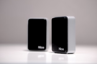 Two rectangular black and silver devices with the word 'Nice' appear on a smooth white surface. They are positioned in such a way that one is in the foreground while the other is slightly blurred in the background.