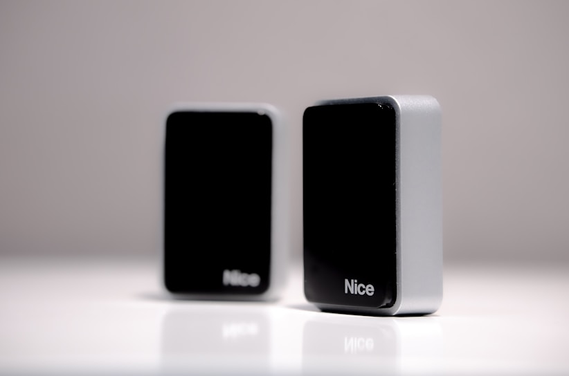 Two rectangular black and silver devices with the word 'Nice' appear on a smooth white surface. They are positioned in such a way that one is in the foreground while the other is slightly blurred in the background.