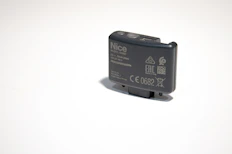 A small, rectangular electronic device with various labels and symbols, including certification marks and model information. The device appears to be made of dark plastic and has a button on top.