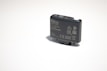 A small, rectangular electronic device with various labels and symbols, including certification marks and model information. The device appears to be made of dark plastic and has a button on top.