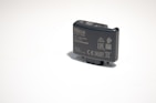 A small, rectangular electronic device with various labels and symbols, including certification marks and model information. The device appears to be made of dark plastic and has a button on top.