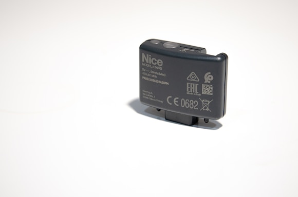 A small, rectangular electronic device with various labels and symbols, including certification marks and model information. The device appears to be made of dark plastic and has a button on top.