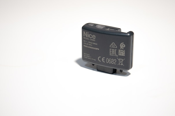 A small, rectangular electronic device with various labels and symbols, including certification marks and model information. The device appears to be made of dark plastic and has a button on top.