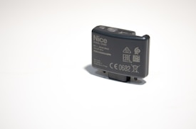 A small, rectangular electronic device with various labels and symbols, including certification marks and model information. The device appears to be made of dark plastic and has a button on top.