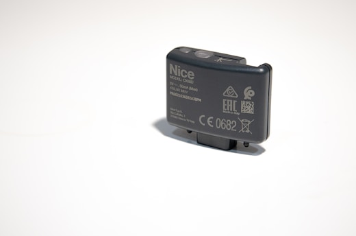 A small, rectangular electronic device with various labels and symbols, including certification marks and model information. The device appears to be made of dark plastic and has a button on top.
