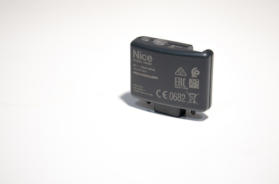 A small, rectangular electronic device with various labels and symbols, including certification marks and model information. The device appears to be made of dark plastic and has a button on top.