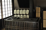 A traditional Japanese interior features a wooden desk area with multiple hanging calligraphy scrolls displaying Japanese characters. The setting includes shoji screens that allow light to filter through, creating a calm and peaceful atmosphere. The desk has an abacus and a box, adding to the vintage aesthetic.