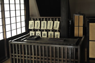 A traditional Japanese interior features a wooden desk area with multiple hanging calligraphy scrolls displaying Japanese characters. The setting includes shoji screens that allow light to filter through, creating a calm and peaceful atmosphere. The desk has an abacus and a box, adding to the vintage aesthetic.