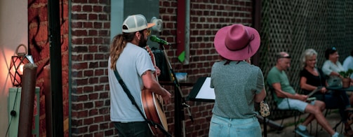 A man and a woman are performing music outdoors. The man is playing a guitar and singing into a microphone, while the woman stands beside him looking at a music stand. They are on a small stage set against a brick wall, and there is a small audience sitting outdoors, watching them. The scene appears relaxed and informal.