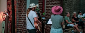 A man and a woman are performing music outdoors. The man is playing a guitar and singing into a microphone, while the woman stands beside him looking at a music stand. They are on a small stage set against a brick wall, and there is a small audience sitting outdoors, watching them. The scene appears relaxed and informal.