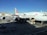 An Asiana Airlines aircraft is stationed at an airport gate. The airplane is undergoing maintenance or loading, with equipment and vehicles around it. Several ground support trucks and cargo loaders are positioned beside the aircraft. The sky is clear, and nearby are buildings and other parked aircraft.