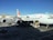 An Asiana Airlines aircraft is stationed at an airport gate. The airplane is undergoing maintenance or loading, with equipment and vehicles around it. Several ground support trucks and cargo loaders are positioned beside the aircraft. The sky is clear, and nearby are buildings and other parked aircraft.