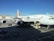 An Asiana Airlines aircraft is stationed at an airport gate. The airplane is undergoing maintenance or loading, with equipment and vehicles around it. Several ground support trucks and cargo loaders are positioned beside the aircraft. The sky is clear, and nearby are buildings and other parked aircraft.