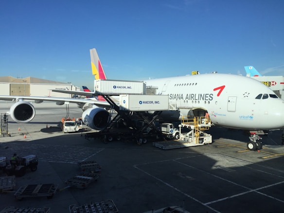 An Asiana Airlines aircraft is stationed at an airport gate. The airplane is undergoing maintenance or loading, with equipment and vehicles around it. Several ground support trucks and cargo loaders are positioned beside the aircraft. The sky is clear, and nearby are buildings and other parked aircraft.