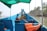 A blue boat with seating and a green canopy, equipped with life jackets and ropes. The boat is docked on a calm waterway with a view of palm trees and a coastal landscape in the background.
