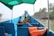 A blue boat with seating and a green canopy, equipped with life jackets and ropes. The boat is docked on a calm waterway with a view of palm trees and a coastal landscape in the background.