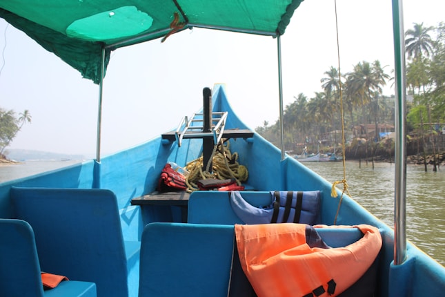 A blue boat with seating and a green canopy, equipped with life jackets and ropes. The boat is docked on a calm waterway with a view of palm trees and a coastal landscape in the background.