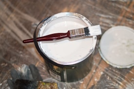 An open can of white paint with a paintbrush resting across the top, set on a surface covered with protective plastic sheeting. The brush has a red handle, and there is a black rubber glove and another brush nearby.