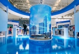 A modern exhibition space features a large cylindrical digital display showing a cityscape with skyscrapers. Surrounding this are several booths with people interacting, some looking at screens and technology displays. The space is well-lit with a sleek, contemporary design using blue and white colors.