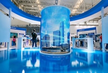 A modern exhibition space features a large cylindrical digital display showing a cityscape with skyscrapers. Surrounding this are several booths with people interacting, some looking at screens and technology displays. The space is well-lit with a sleek, contemporary design using blue and white colors.