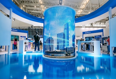 A modern exhibition space features a large cylindrical digital display showing a cityscape with skyscrapers. Surrounding this are several booths with people interacting, some looking at screens and technology displays. The space is well-lit with a sleek, contemporary design using blue and white colors.