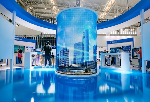 A modern exhibition space features a large cylindrical digital display showing a cityscape with skyscrapers. Surrounding this are several booths with people interacting, some looking at screens and technology displays. The space is well-lit with a sleek, contemporary design using blue and white colors.