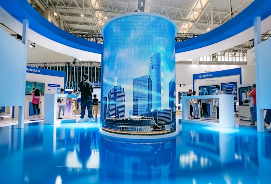 A modern exhibition space features a large cylindrical digital display showing a cityscape with skyscrapers. Surrounding this are several booths with people interacting, some looking at screens and technology displays. The space is well-lit with a sleek, contemporary design using blue and white colors.