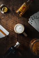 A tabletop scene includes a foamy glass of beer at the center, surrounded by a bottle of whiskey, a small bottle of dark liquor, a slice of lemon, playing cards, and a jar of chips. The setting is rustic, with a wooden surface creating a cozy and casual atmosphere.