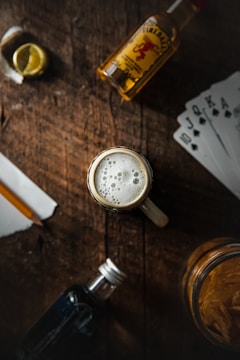 A tabletop scene includes a foamy glass of beer at the center, surrounded by a bottle of whiskey, a small bottle of dark liquor, a slice of lemon, playing cards, and a jar of chips. The setting is rustic, with a wooden surface creating a cozy and casual atmosphere.