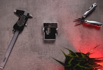 A digital caliper, a small mechanical part, and a multi-tool are arranged on a textured surface. The image also includes some green plant leaves at the bottom right corner with a red light casting a soft glow.